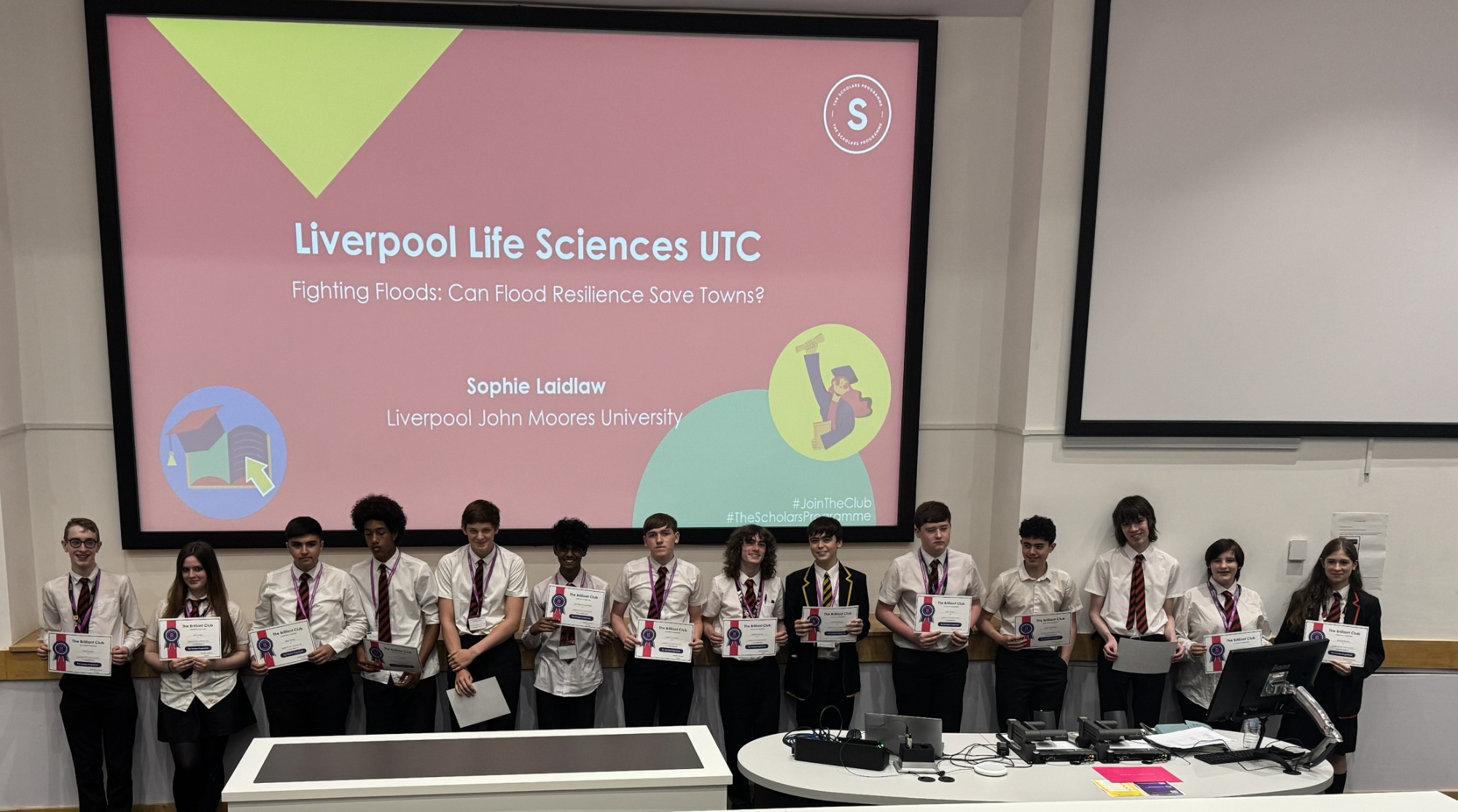Image of scholar programme students stood at the front of a lecture hall receiving their certificates from the Brilliant Club.